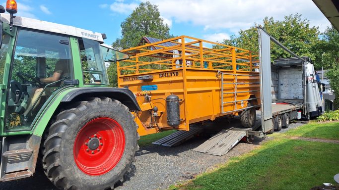 Ein orangefarbener Anhänger wird von einem Traktor und Lkw auf einer Auffahrt transportiert.