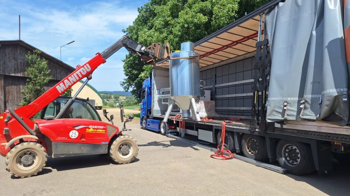 Roter Telehandler entlädt große silberne Behälter von einem Lkw auf einem Bauernhof.